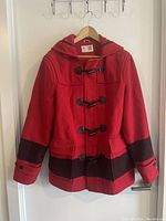 Front view of the red Hudson’s Bay HBC men's coat with black horizontal stripes and toggle closures on a wooden hanger.