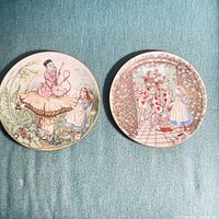 Pair of Limoges porcelain plates with Alice in Wonderland scenes, laid on a blue fabric background.