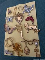 Photo showing 16 various vintage costume jewelry brooches and pins on a yellow backing with blue fabric background. Includes butterflies, heart shapes, animals, flower, ladder, and earrings.