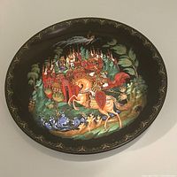Full front view of the round black handpainted collector plate featuring a detailed Russian fairytale scene with knight, castle, and river.