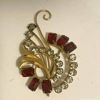 Front view showing gold toned metal brooch with clear and red rhinestones arranged in floral swirl design.