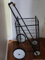 Photo shows black metal shopping cart upright with white wheels and wire basket design.