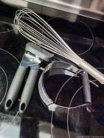 Set of three kitchen utensils on a black glass stovetop: manual can opener, wire whisk, and pastry blender.