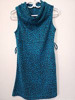 Front view of sleeveless turquoise dress with black leopard print pattern hung on a hanger, showing full length and side ties.