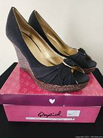 Side view of black open-toe wedge shoes on top of pink branded box showing the full profile and wedge heel.