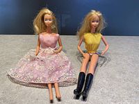 Two vintage Barbie dolls seated, one in a pink dress with floral lace and the other in a yellow dress with black boots.