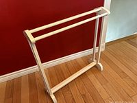 Full view of antique white painted quilt stand blanket rack against a red wall and hardwood floor showing the two horizontal bars and side supports with curved base feet.