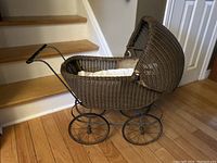 Side view of antique wicker baby doll toy stroller showing wicker weaving, curved hood, metal wheels, and taped handle repair.
