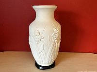 Front view of white bisque pottery vase with raised daffodil and leaf relief pattern and black lacquer base against red background.