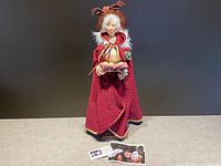 Full standing view of the Jacqueline Kent Old Woman doll wearing a red cloak and holding a basket, placed on a flat surface with a neutral background.