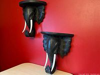 Pair of black, carved wooden elephant head wall shelves mounted on a red wall, each with white tusks and oval shelf tops, shown from angled front views.