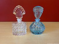 Front view of two vintage pressed glass perfume bottles with blown glass stoppers, one clear and one turquoise blue, on a wooden surface against a red background.