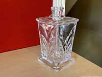 Clear square crystal ice bucket with leaf pattern and matching lid shown on wooden surface against red wall.