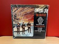 Full front view of The Beatles 'Something New' album cover in plastic sleeve against red background on wooden surface.
