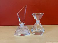Pair of crystal perfume bottles on wood surface against red wall, showing geometric stopper shapes and bases.