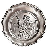 Front view of vintage pewter plate showing embossed lady spinning wool in a kitchen scene