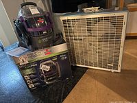 Bissell SpotClean Pet Pro cleaner, its box, and electric fan shown side by side on floor.