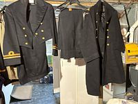 Photo showing two navy wool double-breasted jackets with gold brass buttons and two pairs of matching navy wool trousers hanging on a rack.