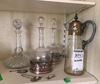 Three clear glass decanters on shelf, two with metal holders and tags, one with metal lid and handle.