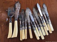 Photo showing set of serving fish forks and cutlery with cream-colored handles and silver collars, laid out in a row on wooden surface.