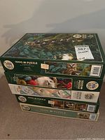 Lot of four Cobble Hill puzzle boxes stacked on floor, showing the sides and fronts with puzzle artwork and labels.