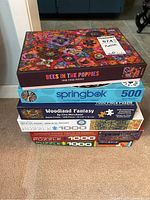 Stack of seven jigsaw puzzle boxes showing various brands and piece counts, the top one "Bees in the Poppies", 1000 pieces by Galison.