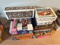 Stack of assorted jigsaw puzzles including Eurographics, Beach Cat, Autumn Harvest and others visible with colorful boxes and puzzle piece counts labeled mostly 1000.
