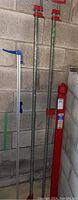 Two 6-foot pipe clamps with visible red handles next to a 5-foot aluminum/plastic clamp with blue handle, all standing against a concrete block wall.