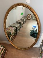 Front view of the oval mirror resting against a wall, showing the full gilt frame and reflective surface with visible scratches and some background reflection in the glass.