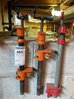 Three pipe clamps in orange and red mounted on 16-inch metal pipes, shown against a basement wall.