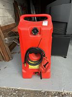 Front view of the red portable gas tank with black nozzle and hose coiled at the front, standing on the floor near wooden planks and other stored items.