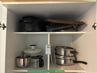 Upper cabinet shelf with 3 black enamel roasters with lids on left side and 2 stainless steel lidded pots and white coffee carafe on right side.
