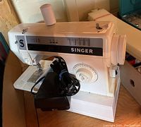 Front left side angle of the white Singer DX-14C sewing machine with spool and foot pedal showing power cord wrapped.
