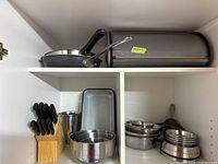 Photo of two kitchen shelves with various items: wooden cutlery block, stainless steel pet bowls stacked, metal bread box, enameled container, two skillets with handles, and baking tray