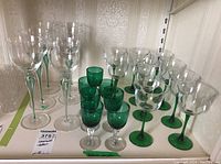 Group of 23 stemware glasses arranged on a shelf showing different styles including green and clear glasses with clear and green stems.