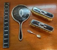 Top view of four-piece sterling silver vanity set with ruler for scale. Items include a mirror, two brushes, and a small spoon all featuring silver decoration and dark inlays.