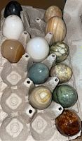 Top view showing 12 assorted polished stone eggs arranged in a cardboard egg carton with visible natural patterns and colors ranging from black, white, green, beige, and brown.