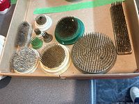 Photo showing 10 wire flower pin frogs arranged in a cardboard box. Various sizes and base materials including metal and plastic. Close view of spikes and bases.