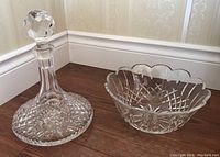 Photo showing Waterford crystal decanter with faceted stopper and crystal bowl with scalloped edges and intricate cut glass pattern on wooden floor.