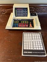 Front and top view of electronic handheld game with control buttons and screen grid, along with original notepad placed in front