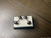 Pair of oval-shaped sterling silver cuff links resting in an open small jewelry box with white padding background on a wooden surface