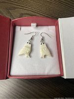 Pair of white carved owl earrings in red jewelry box on a wood surface.