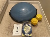 Blue Bosu Ball half dome balance trainer with two yellow resistance bands and instruction manual on beige carpet.