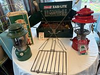 Photo of green and red Coleman lanterns, Coleman Easi-Lite camping stove, metal grilling rack, and fork accessories arranged on a white table.