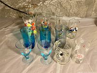 Wide view showing assortment of colorful vintage glasses and plastic cups including McDonald's Batman Forever Two-Face glass mug, Snoopy mug, blue tinted glasses, and plastic shooters with geometric designs.
