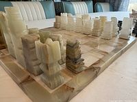 Angled close-up of chess pieces on marble board showing the blocky geometric carved style and various beige tones. Pieces are large and heavy with some chips and wear.