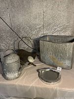 Full lot showing ash can, watering can, ashtray bowl, and firewood carrier on a cloth surface against a stone wall
