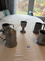 Set of five pewter drinking vessels on a white tablecloth, including a large tankard with lid, two small goblets, and two mugs.