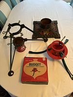 Photo showing three Christmas tree stands arranged on a table: black metal stand, decorative cast iron base, and Budget stand with box and components visible.