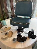 View of folding boat chair with black vinyl seat and back cushion, two silver Mercury propellers, and two black Johnson propellers on table.